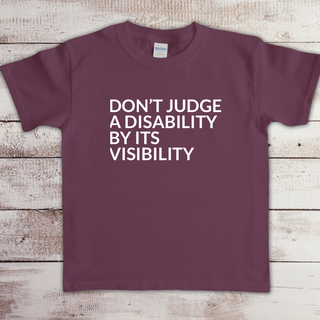 Toddler t-shirt with the phrase “Don’t judge a disability by its visibility” in white vinyl, designed for disability awareness and inclusion. Available in multiple colors, including maroon and blue.